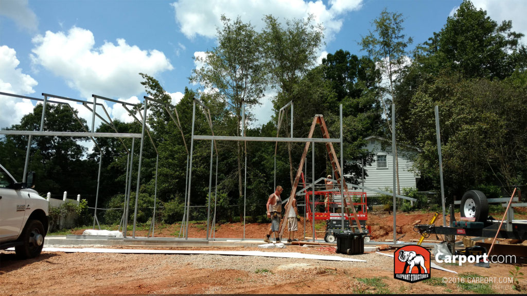 Framing of the building is erected, and two workers use a ladder to assemble the truss system.